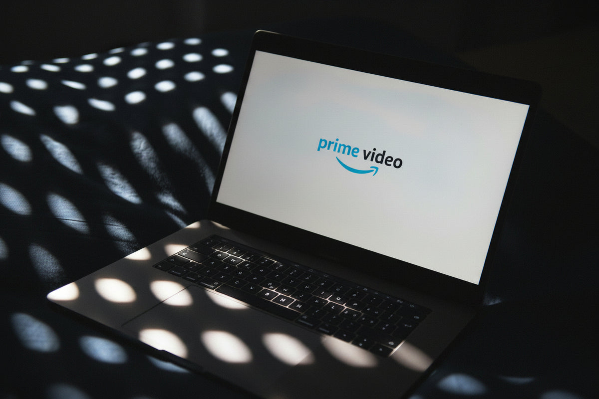 An open laptop on a dark surface with dramatic circular shadows cast across it. The laptop screen displays the Amazon Prime Video logo on a light background.
