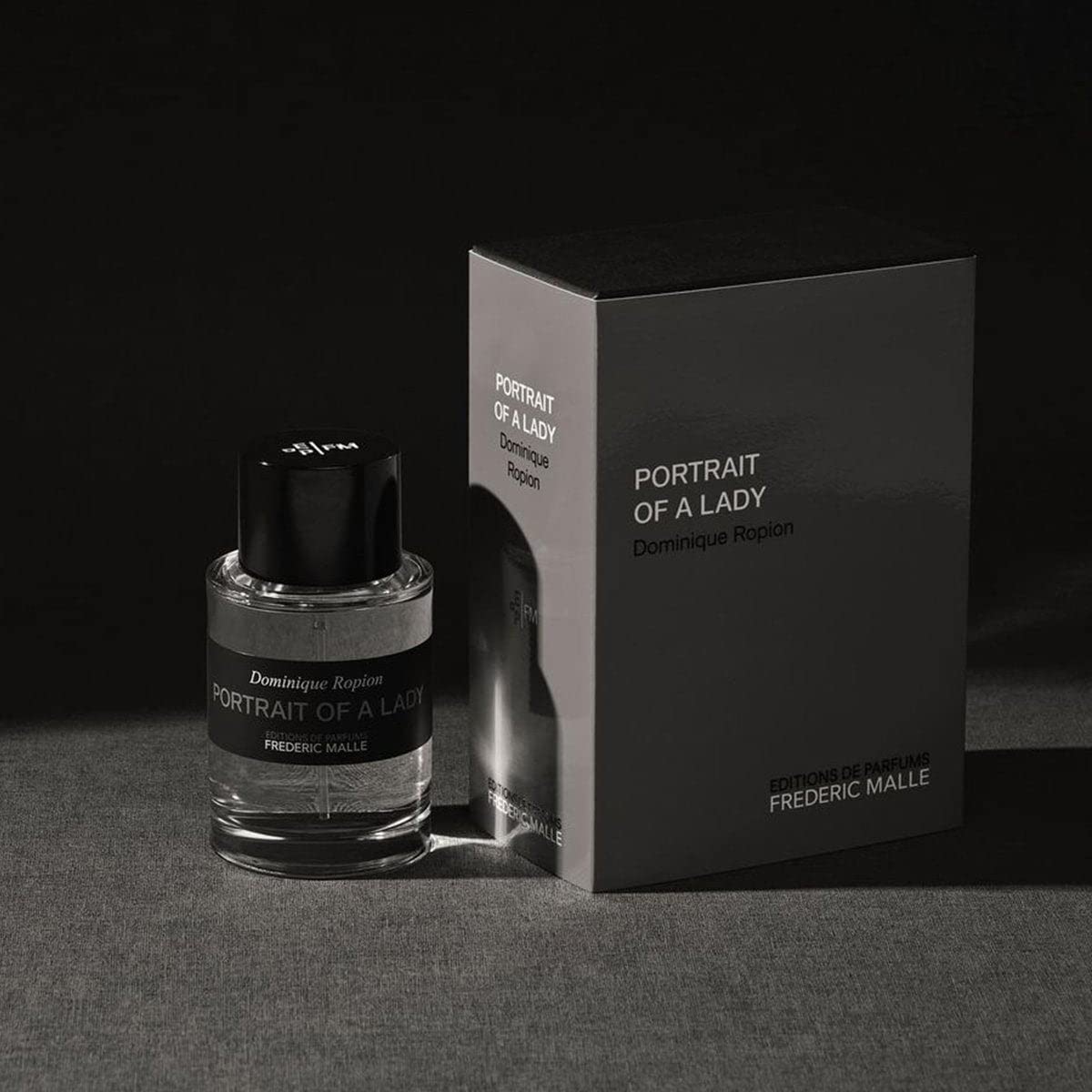Frederic Malle Portrait Of A Lady travel spray bottle (50ml) and its black cylindrical case, presented side-by-side against a dark, textured background
