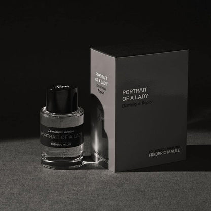 Frederic Malle Portrait Of A Lady travel spray bottle (50ml) and its black cylindrical case, presented side-by-side against a dark, textured background