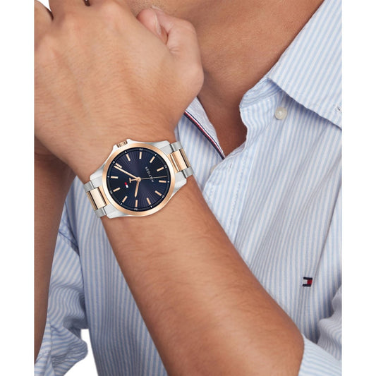 Tommy Hilfiger men's watch with a dark dial and gold-tone case, worn on a wrist against a light blue shirt background.